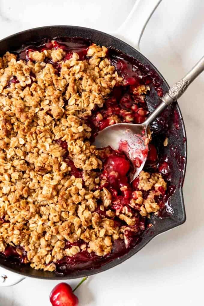 A large serving spoon scooping out a serving of cherry crisp from a cast iron skillet.