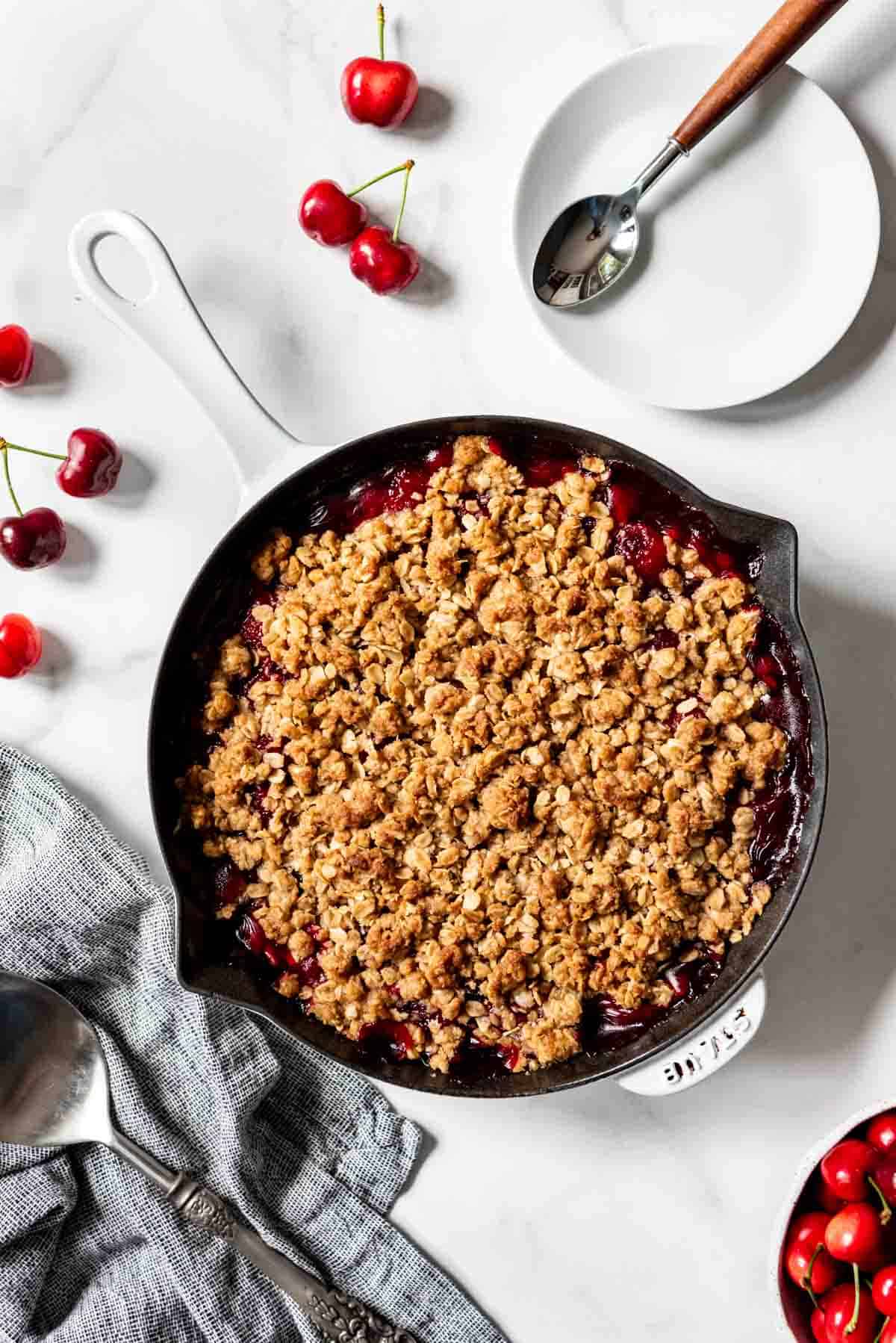 A large cast iron skillet with crisp oat topping over cherry pie filling next to some fresh cherries and a white plate.