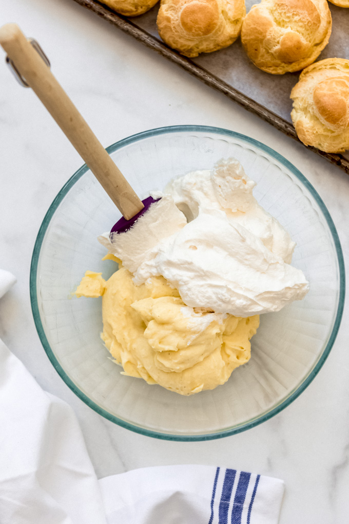 A glass bowl with pastry cream and whipped cream next to cream puff shells.