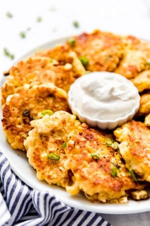 Piles of corn fritters beside a small bowl of sour cream.
