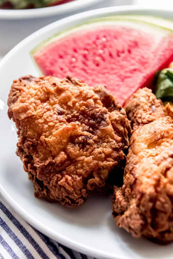 A close image of fried chicken thigh on a plate with sliced watermelon in the background.
