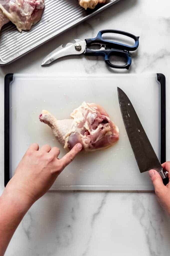 a finger pointing to where to cut to seperate a chicken thigh from a chicken drumstick and another hand holding a knife to the side ready to cut