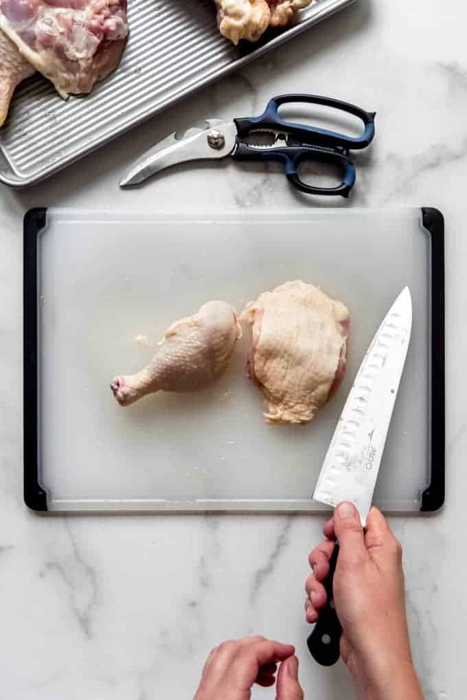 a raw chicken thigh and a chicken drumstick on a cutting board with a pair of hands holding a large knife