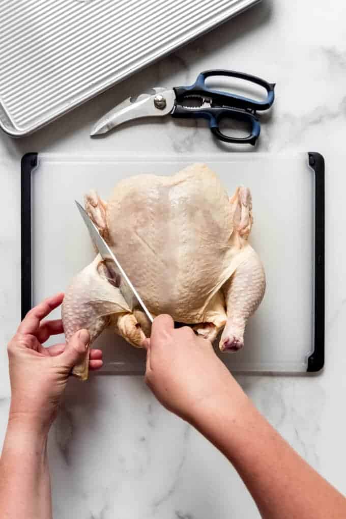 one hand holding a chicken leg and another slicing it off of a whole chicken using a large knife