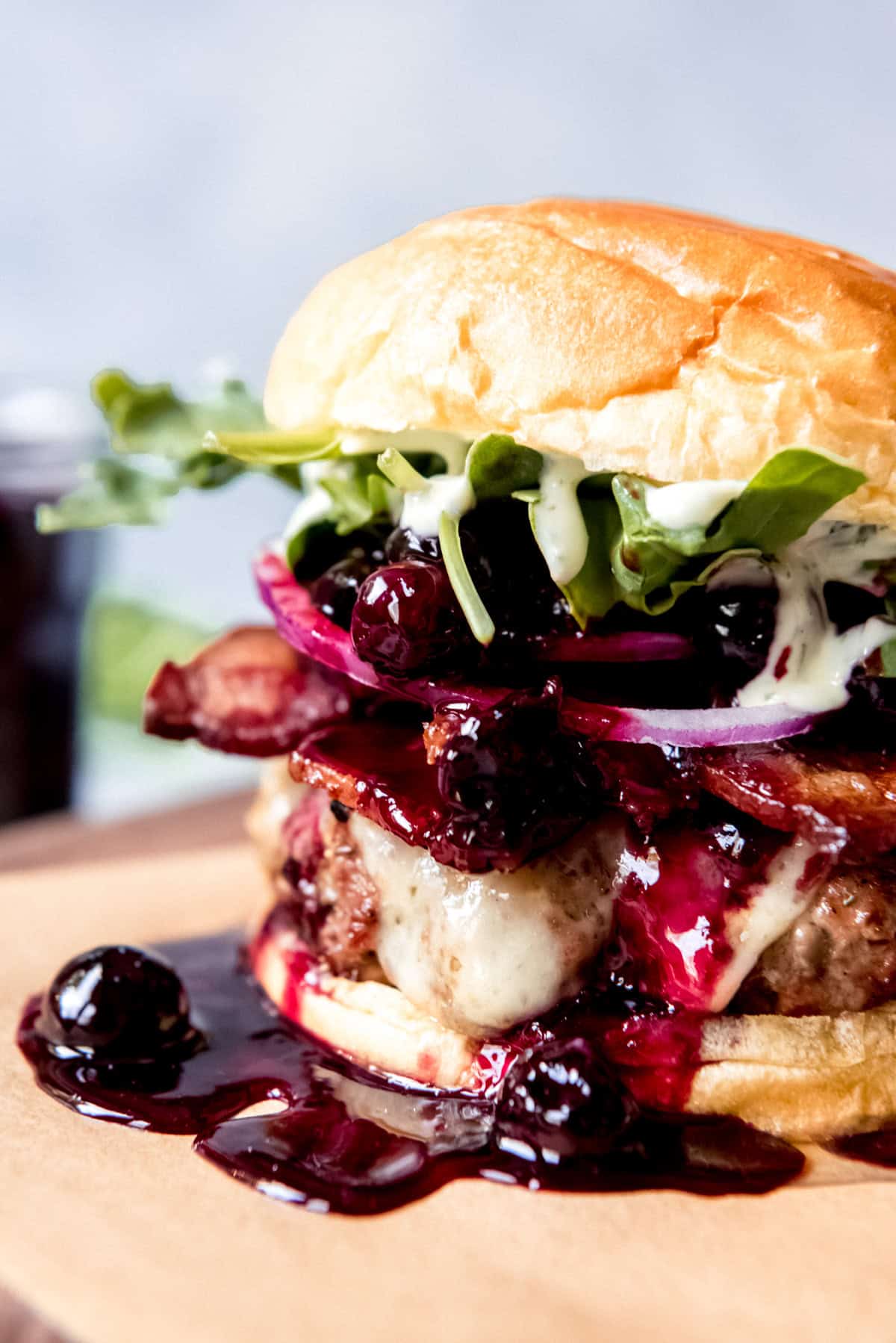 An sideview image of a tall hamburger with cheese, bacon, onions, arugula, basil aioli, and a blueberry balsamic compote.