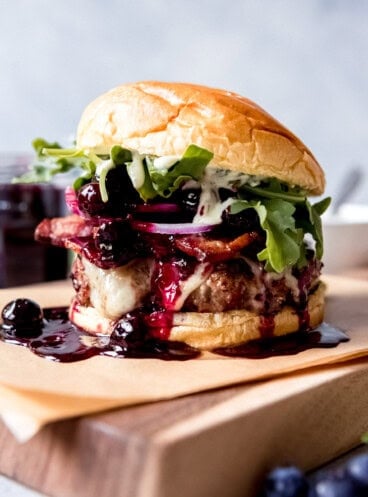 A Red, White, and Blueberry Bacon Burger with Basil Aioli on a piece of parchment paper on top of a wooden cutting board