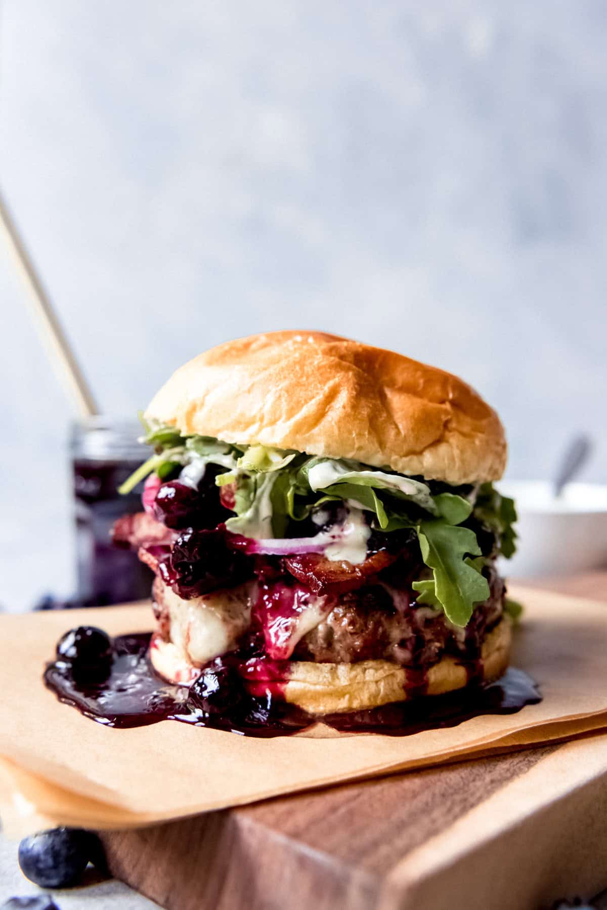 A Red, White, and Blueberry Bacon Burger with Basil Aioli on a piece of parchment paper on top of a wooden cutting board