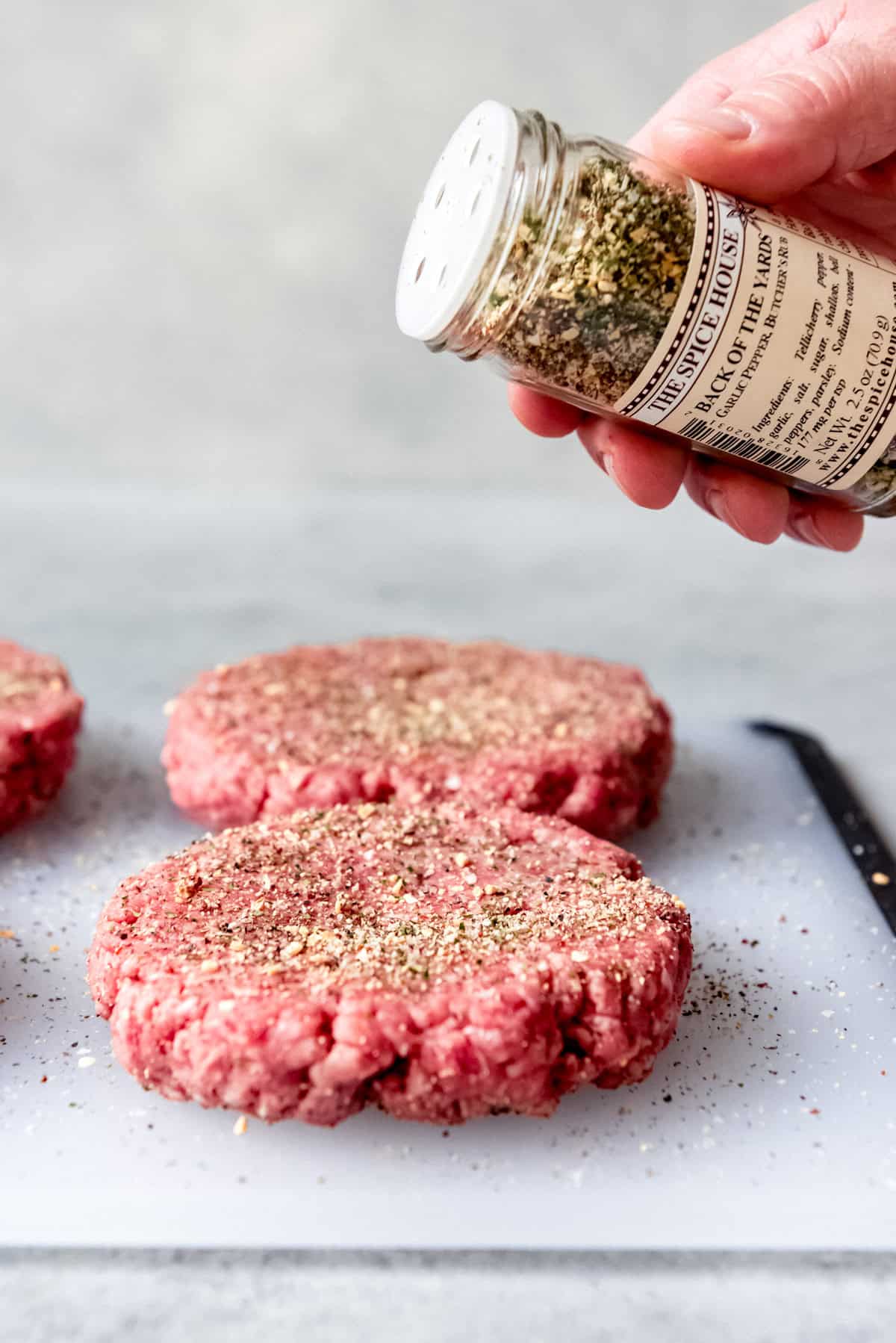 A jar of burger seasoning being held over ground beef patties.