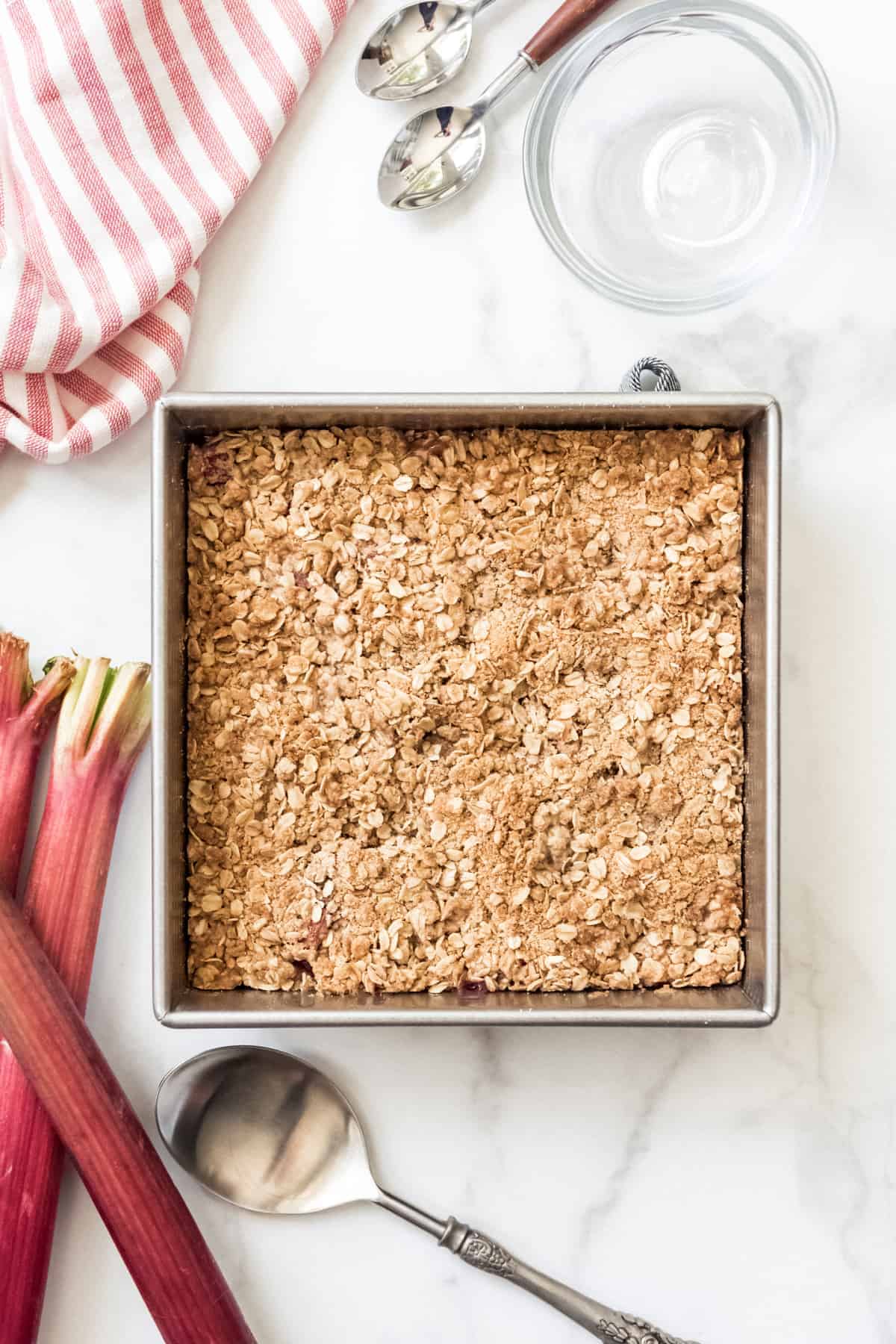 A square baking dish filled with oat crisp topping next to stalks of fresh rhubarb.