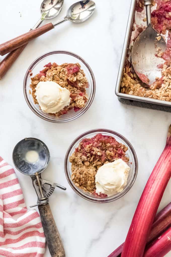 Two glass bowls with rhubarb crisp and scoops of ice cream next to an ice cream scoop and rhubarb stalks.