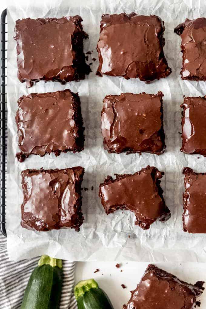 Zucchini brownies with chocolate icing that are cut into squares and spaced apart on parchment paper.