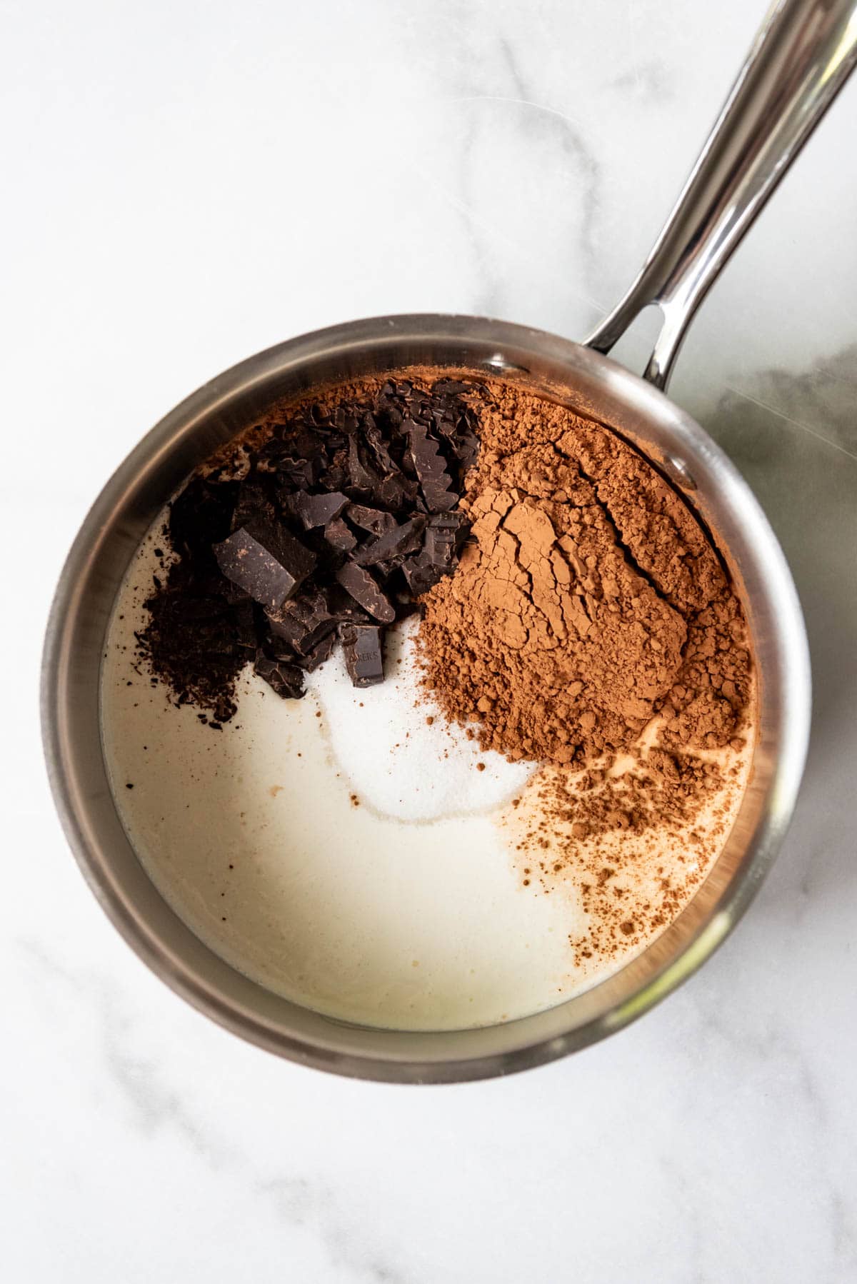 A saucepan with cream, milk, sugar, cocoa powder, and chopped unsweetened chocolate on a marble surface.