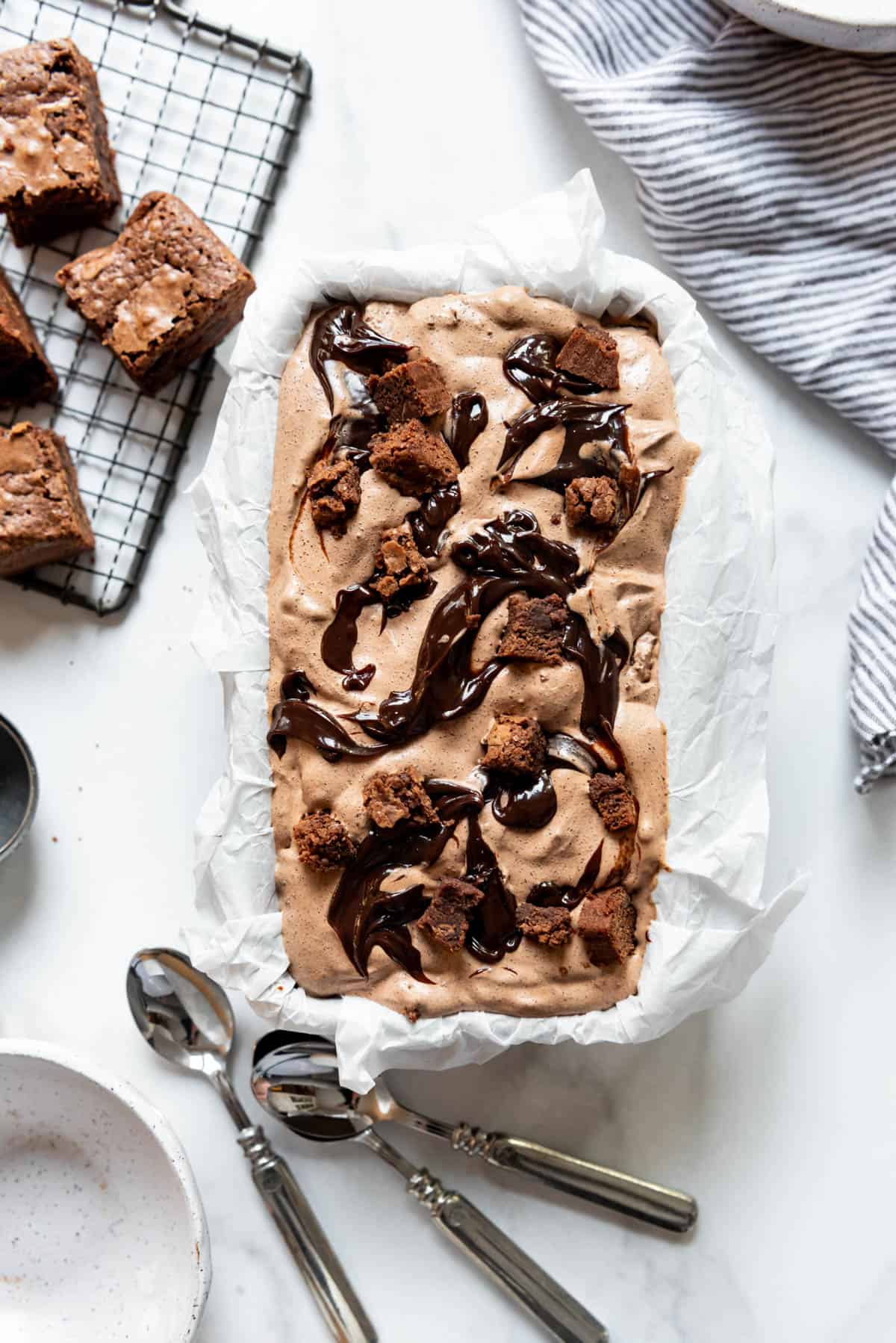 A bread pan filled with homemade chocolate ice cream with brownie pieces and a fudge swirl next to spoons and more brownies.