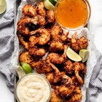 A sheet pan piled with coconut shrimp with lime wedges and two dipping sauces.