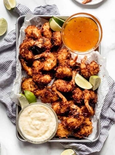 A sheet pan piled with coconut shrimp with lime wedges and two dipping sauces.