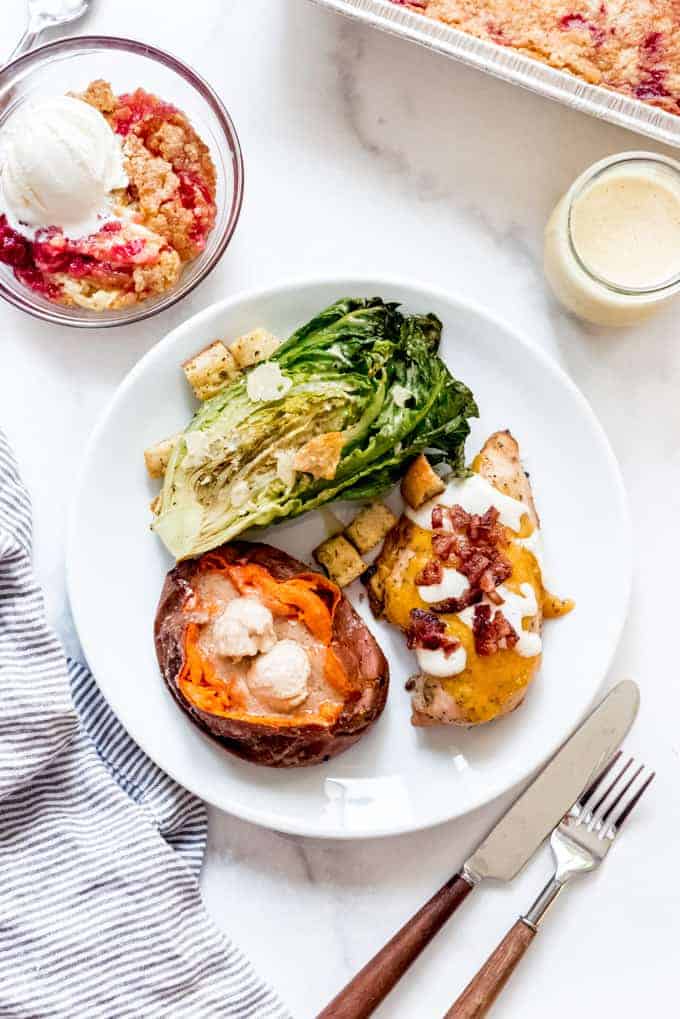 A white plate with a smoked sweet potato with cinnamon honey butter, grilled caesar salad, and bacon ranch chicken next to a striped linen napkin, knife, and fork, with a bowl of cherry pineapple dump cake for dessert.
