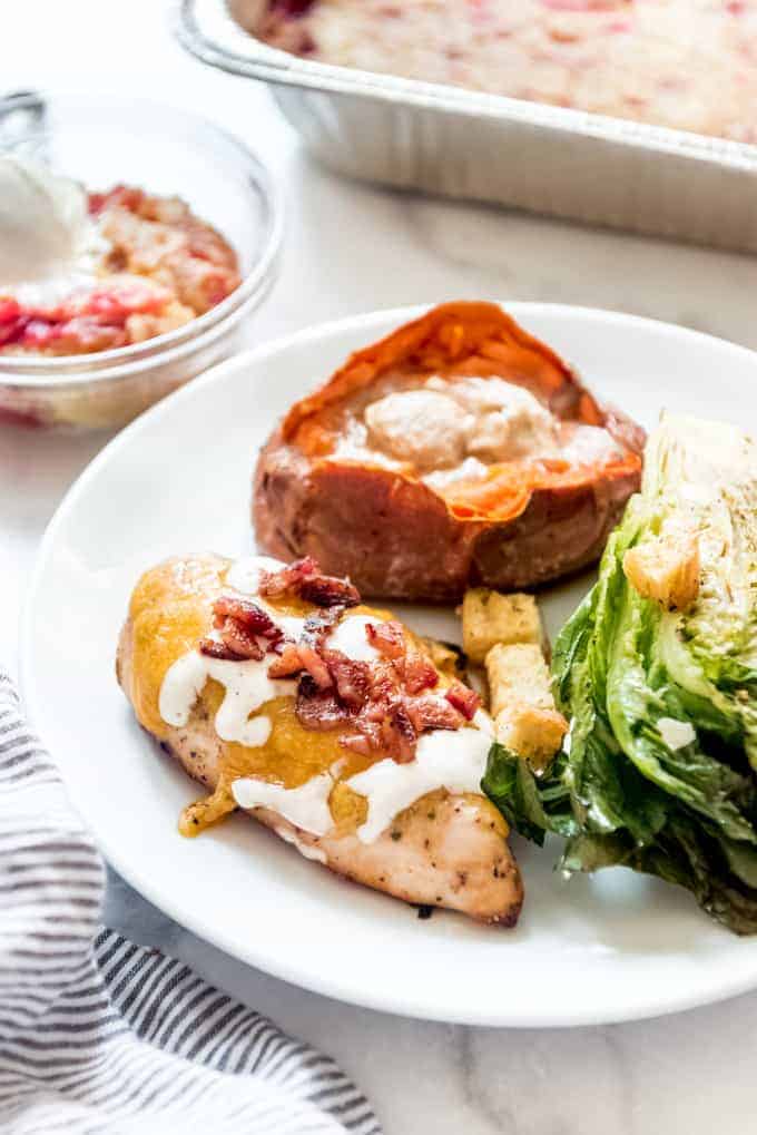 A white plate with bacon ranch chicken, a sweet potato, and grilled romaine lettuce on it next to a bowl of dump cake for dessert.