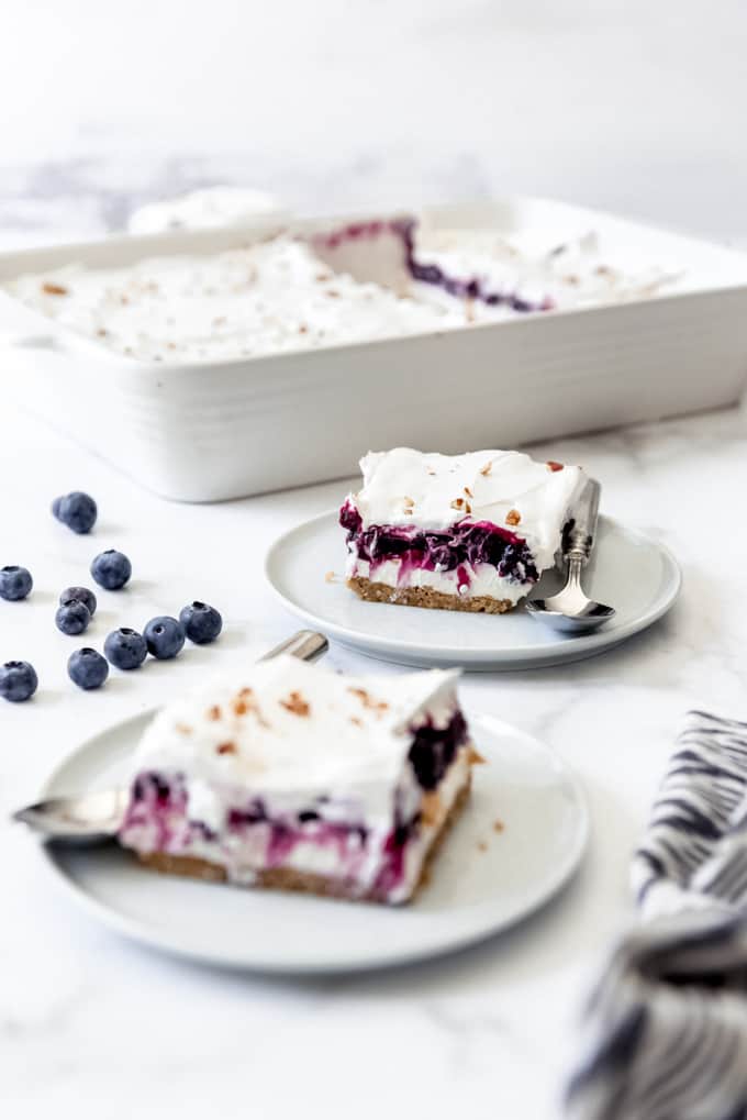 Pieces of blueberry delight on small plates next to fresh blueberries and a white baking dish.