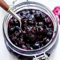 A glass jar of blueberry pie filling with a spoon lifting up some of the filling.