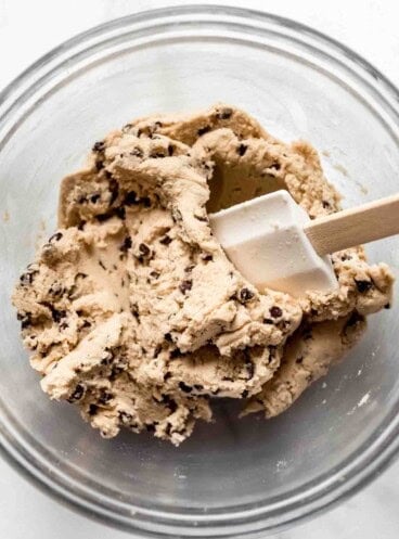 A glass bowl with a white spatula mixing mini chocolate chips into cookie dough.