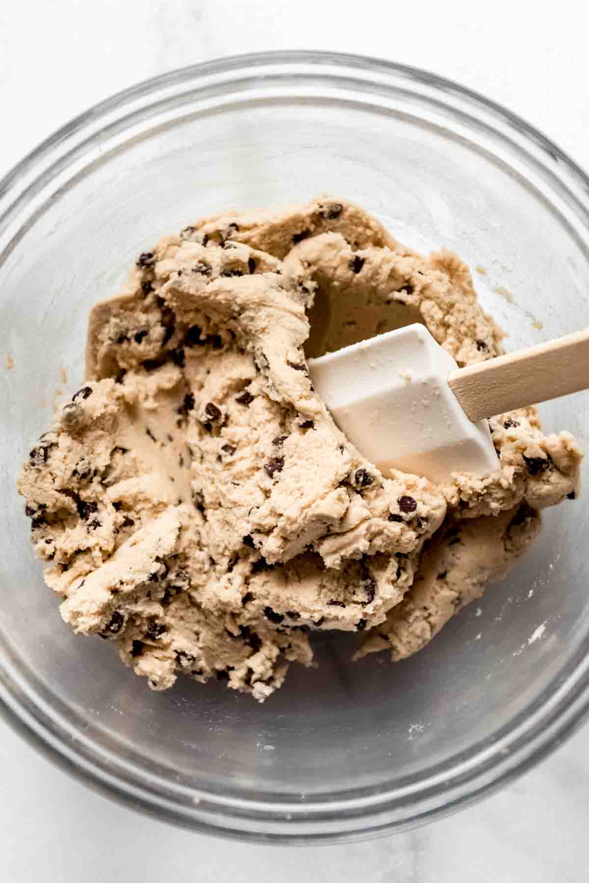 A glass bowl with a white spatula mixing mini chocolate chips into cookie dough.