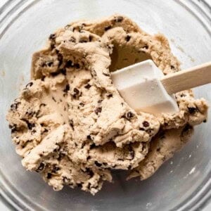 A glass bowl with a white spatula mixing mini chocolate chips into cookie dough.