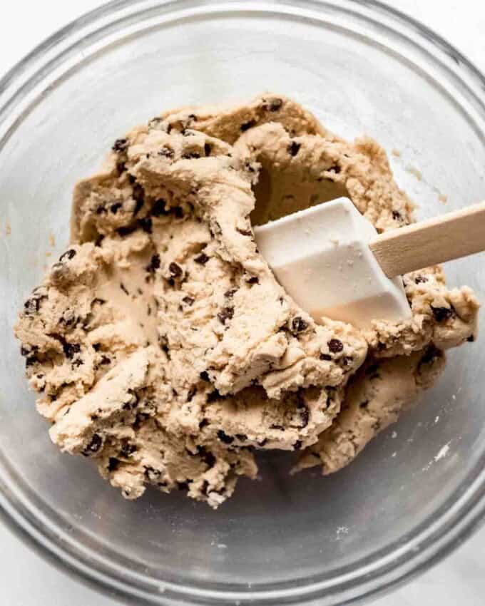 A glass bowl with a white spatula mixing mini chocolate chips into cookie dough.