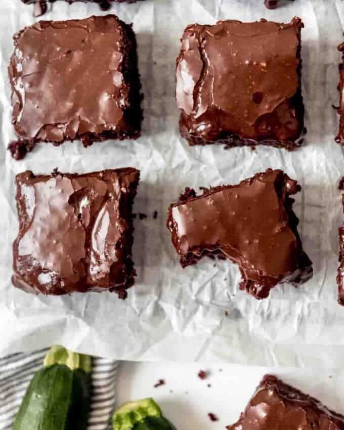 Zucchini brownies with chocolate icing that are cut into squares and spaced apart on parchment paper.