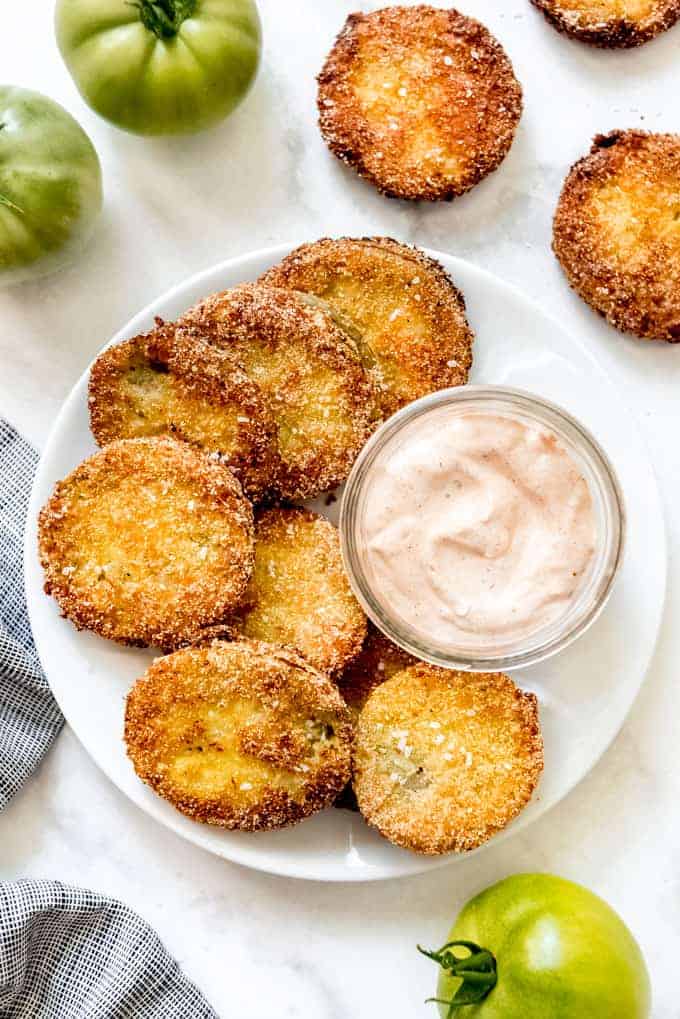 Fried green tomatoes on and off of a white plate with a jar of dipping sauce and whole green tomatoes to the side