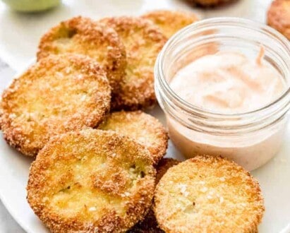 Fried green tomatoes on a white plate with a jar of dipping suace and some whole green tomatoes to the sides