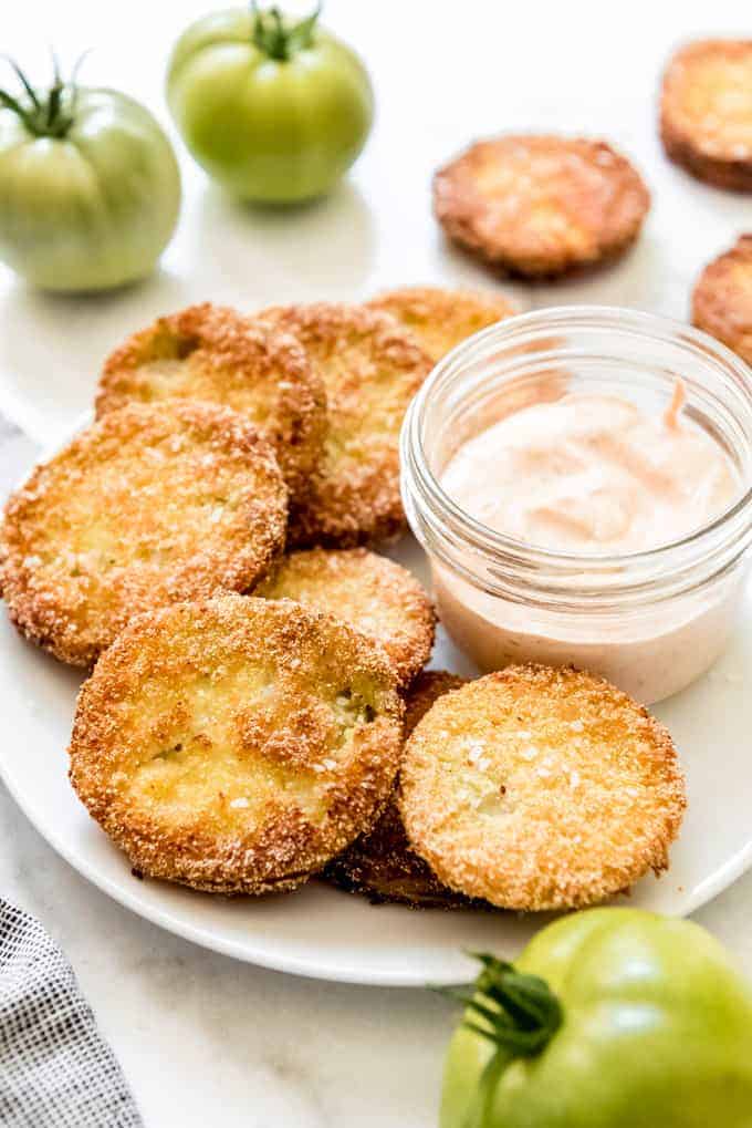 Fried green tomatoes on a white plate with a jar of dipping suace and some whole green tomatoes to the sides