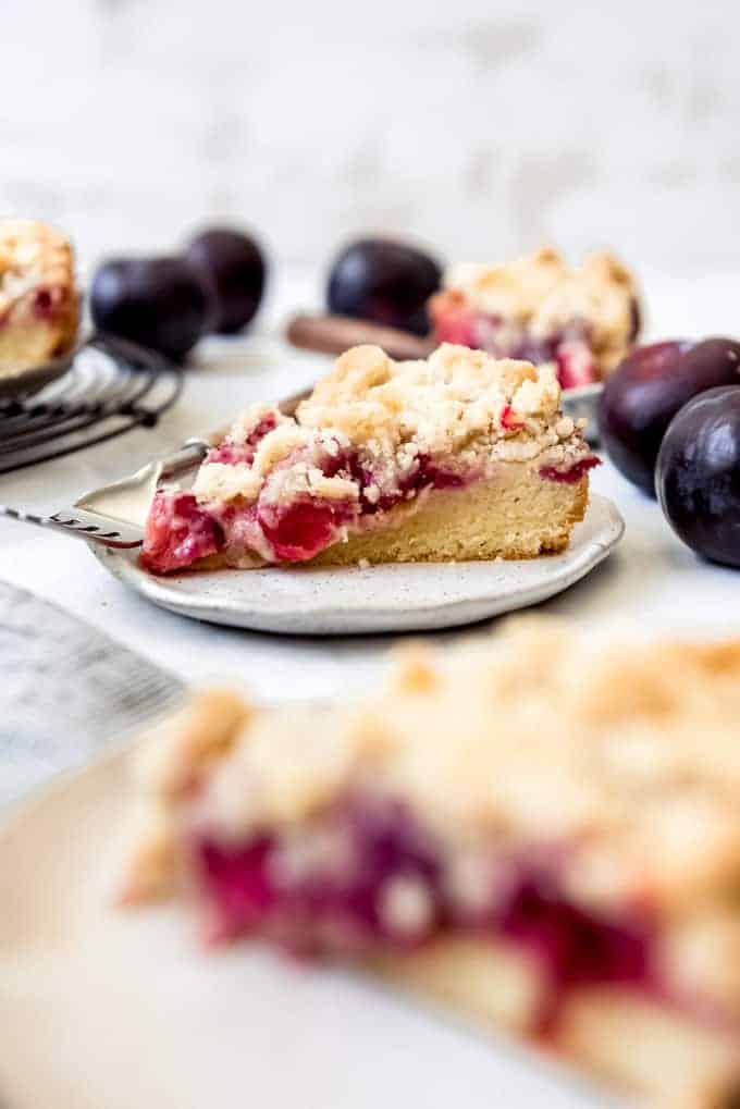 A slice of plum cake with streusel topping on a plate next to fresh plums and more slices of cake.