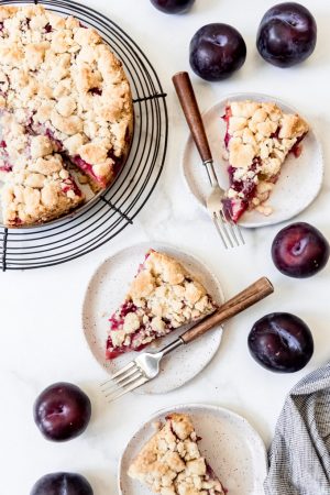 A sliced German plum cake with servings on plates and surrounded by fresh whole plums
