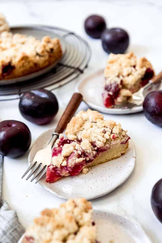 A slice of German plum cake on a speckled plate with a fork, surrounded by dark purple plums.