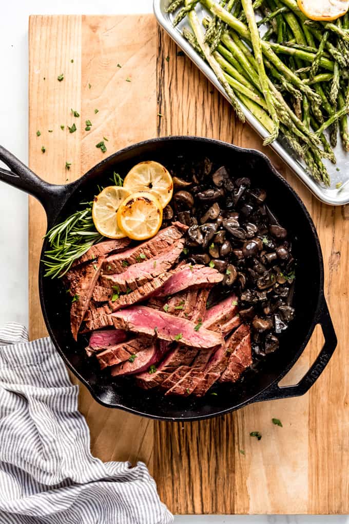 A skillet of sliced flank steak and mushrooms next to roasted asparagus.