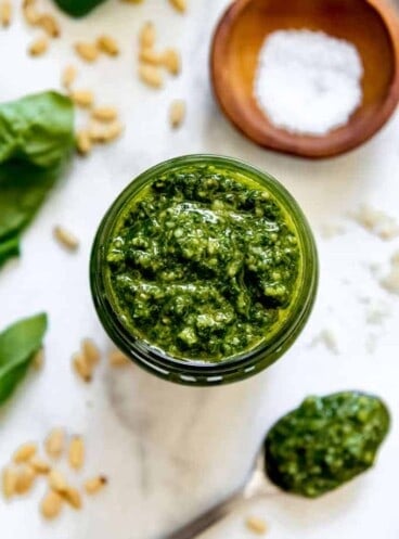 A close image of homemade basil pesto next to a spoonful of pesto, pine nuts, and basil leaves.