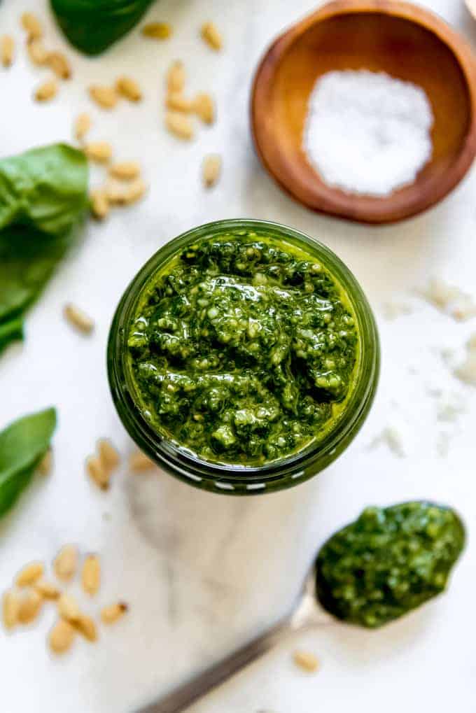 A close image of homemade basil pesto next to a spoonful of pesto, pine nuts, and basil leaves.