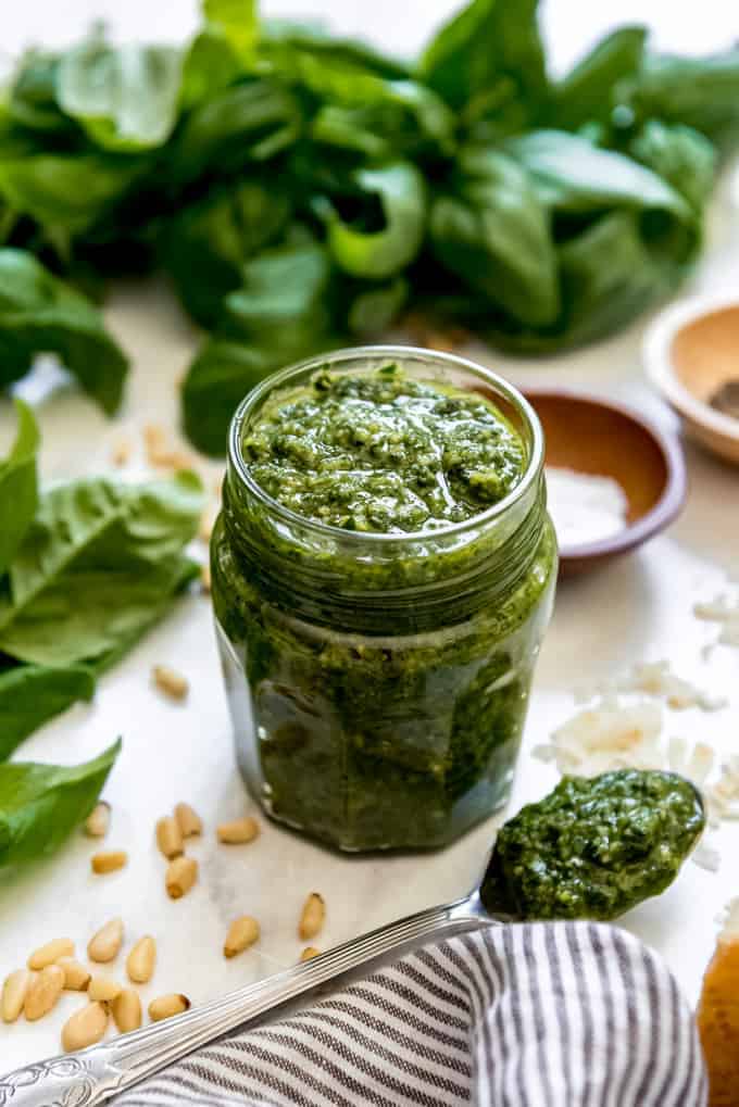A glass jar of homemade basil pesto next to a spoonful of pesto, pine nuts, basil leaves, and a striped cloth napkin.