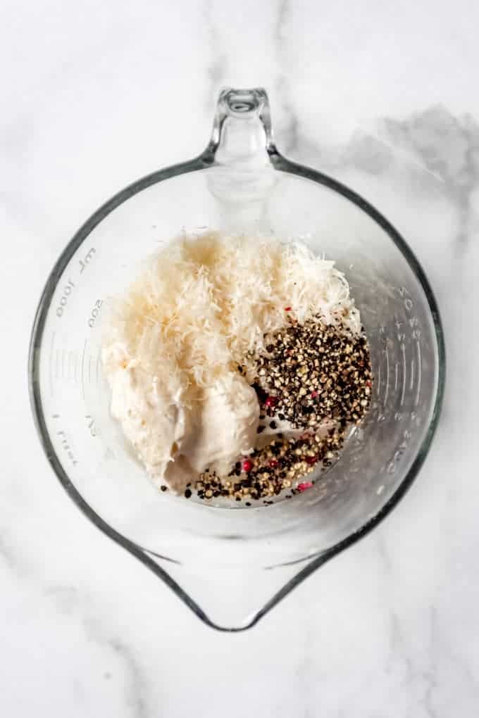 A large mixing bowl with mayo, greek yogurt, grated parmesan cheese, and cracked peppercorns.