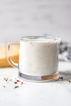 A small glass pitcher filled with creamy parmesan peppercorn dressing with cracked peppercorns and grated parmesan sprinkled nearby.