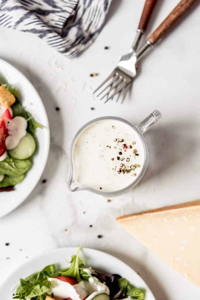 A glass pitcher of parmesan peppercorn dressing with crushed peppercorns sprinkled on top next to two white plates with salad.