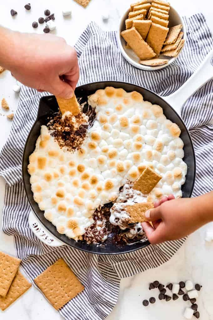 hands scooping melted chocolate and toasted marshmallows out of a skillet with graham crackers