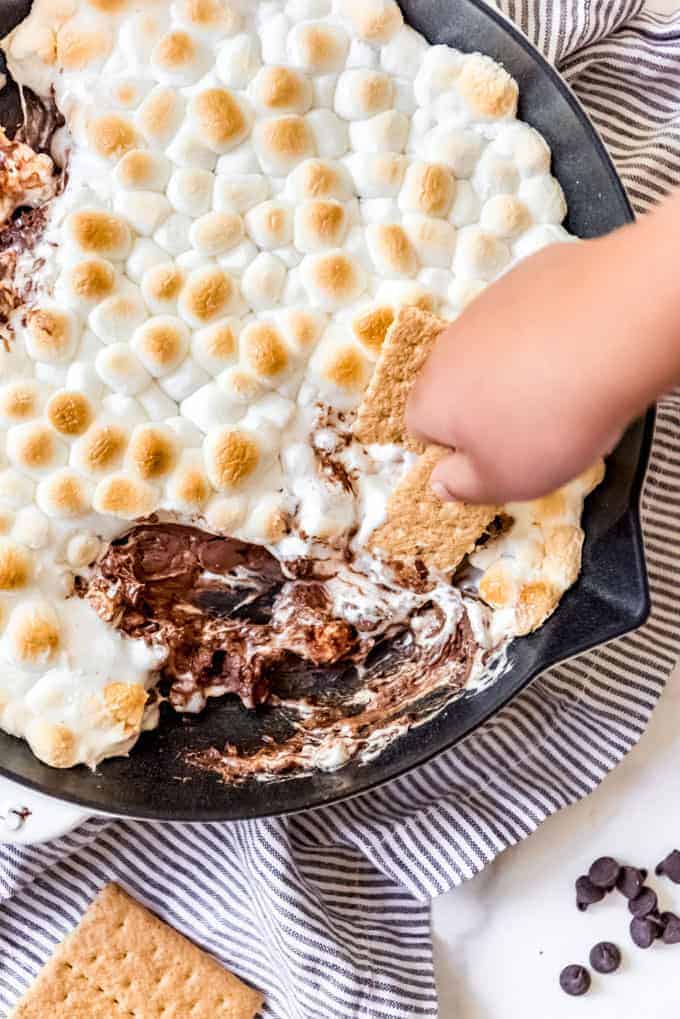 a hand holding a graham cracker to scoop chocolate and marshmallows out of a skillet