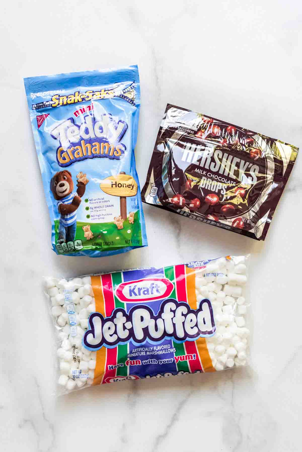 Packages of mini Teddy Grahams, Hershey's milk chocolate drops, and Kraft Jet-Puffed marshmallows on a white marble surface.