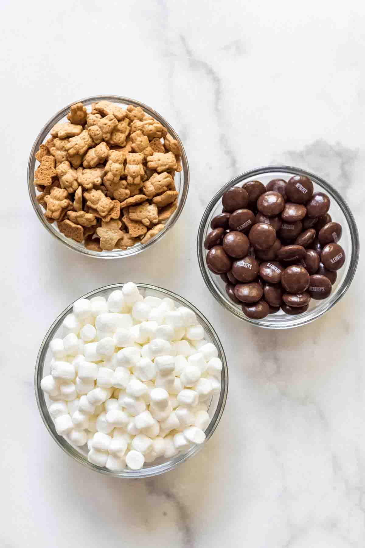 Teddy grahams, Hershey's chocolate drops, and mini marshmallows in three individual bowls on a white marble surface.