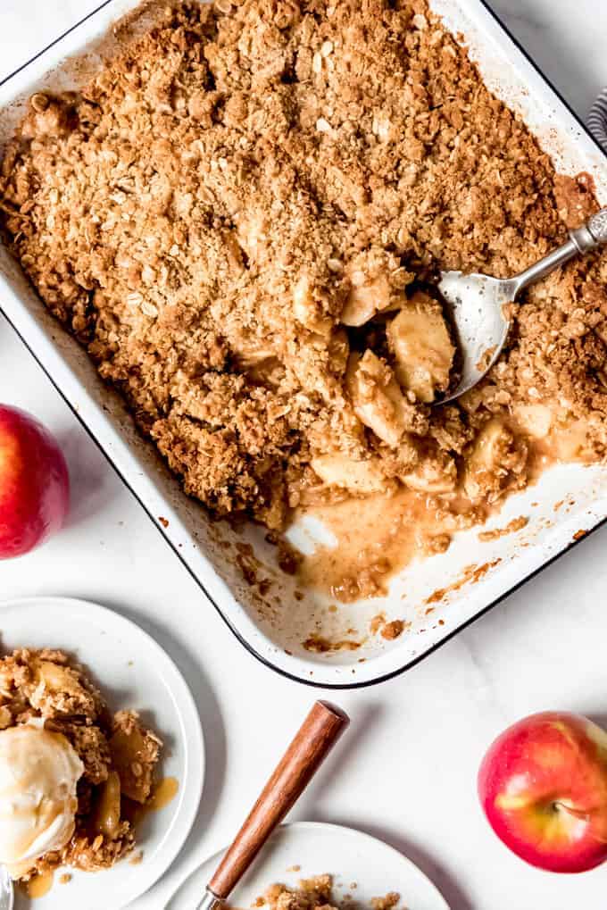 A serving spoon scooping out apple crisp from a baking pan next to plates and apples..