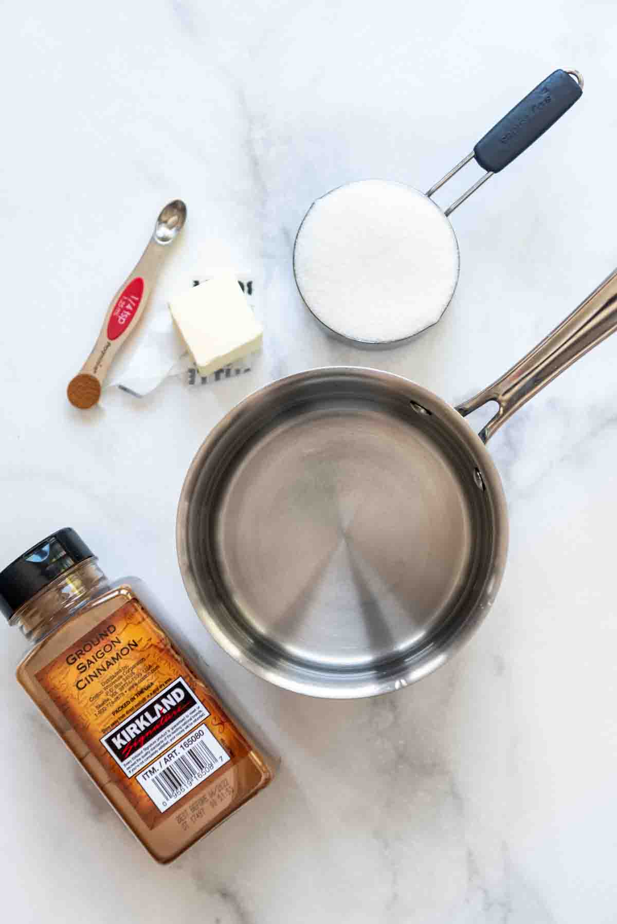 A small pan with cinnamon, sugar, butter, and water on a white marble surface.