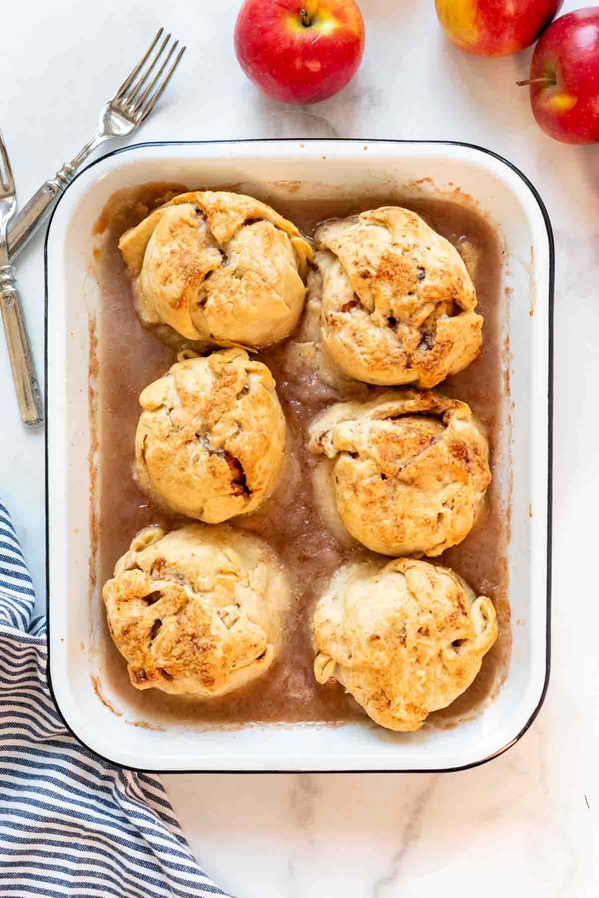 Six apple dumplings in a white enamel baking dish next to red apples.