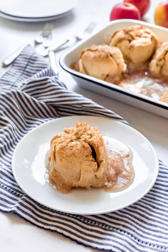 An apple dumpling on a white plate in front of a baking pan with more dumplings.