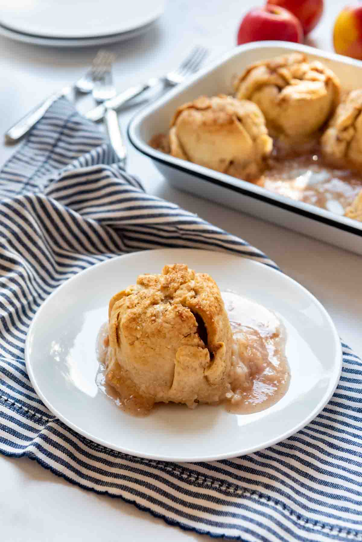 An apple dumpling on a white plate in front of a baking pan with more dumplings.